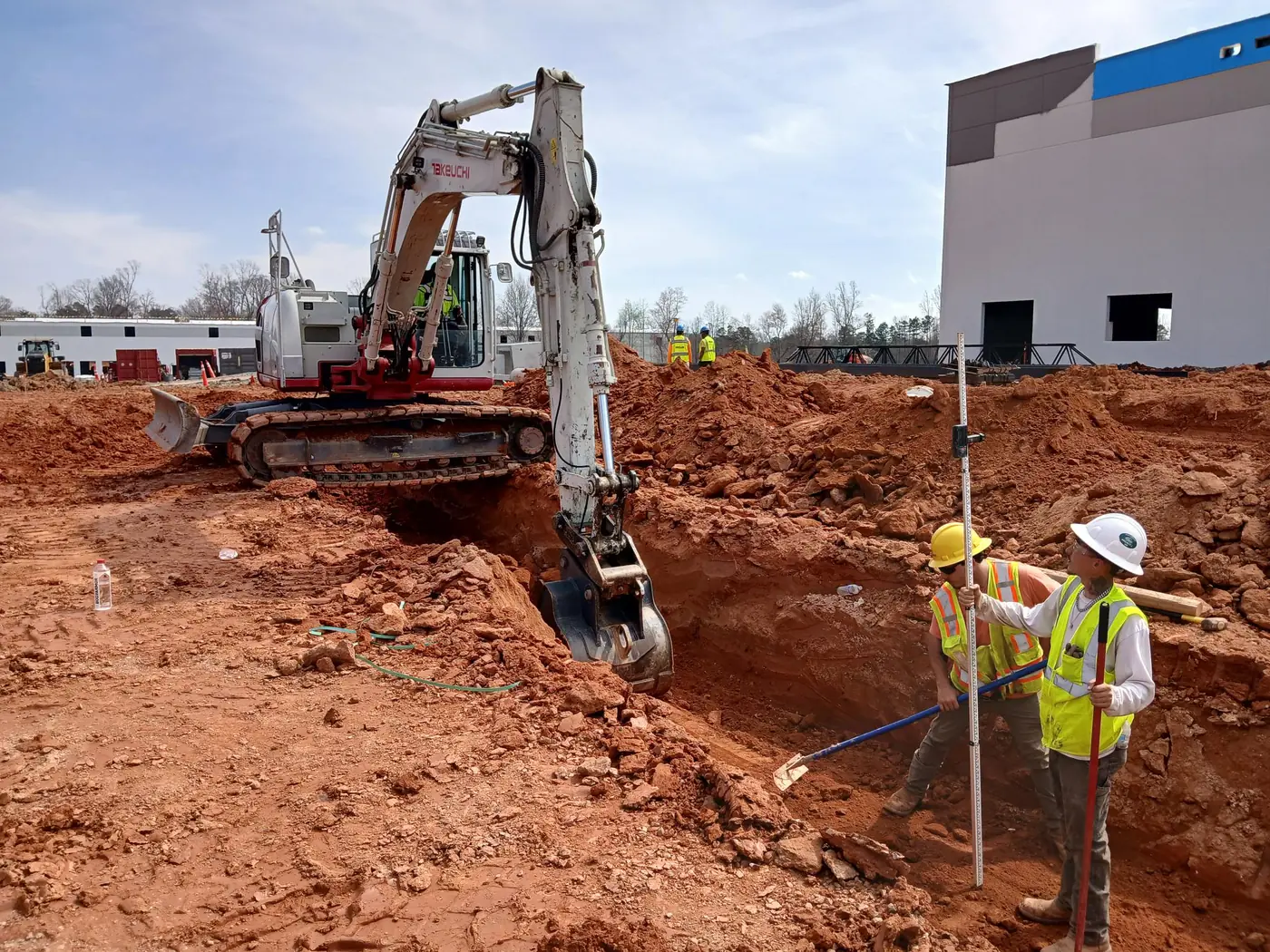 Crew checking trench depth with a grade rod beside an excavator